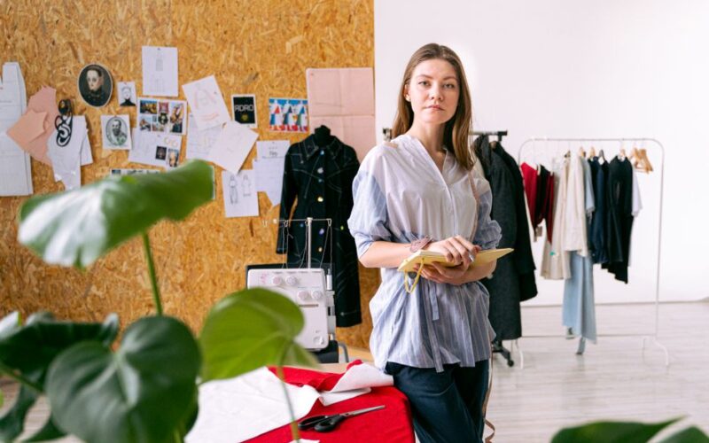 Fashion designer standing in a creative studio surrounded by clothing and sketches.