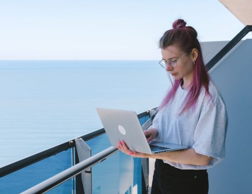 A young woman works remotely on her laptop by the sea on a sunny day, embodying the digital nomad lifestyle.
