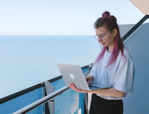 A young woman works remotely on her laptop by the sea on a sunny day, embodying the digital nomad lifestyle.