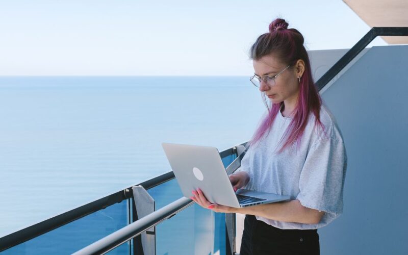 A young woman works remotely on her laptop by the sea on a sunny day, embodying the digital nomad lifestyle.