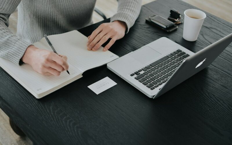 A person in a grey sweater writing in a notebook at a black desk