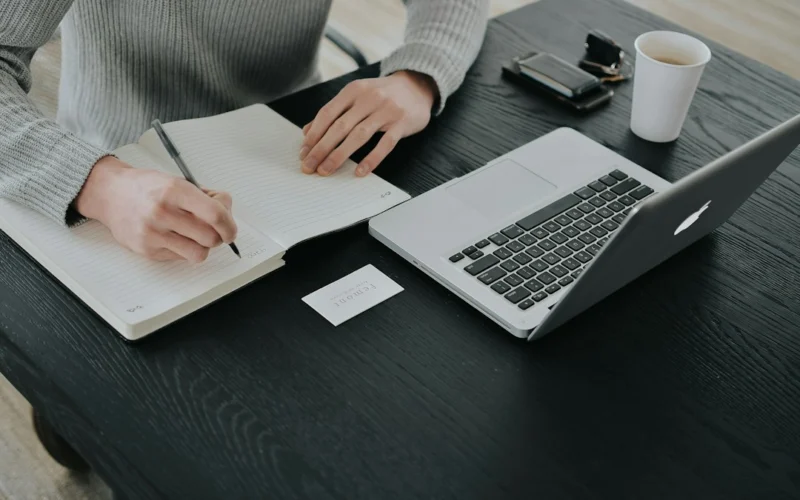 A person in a grey sweater writing in a notebook at a black desk
